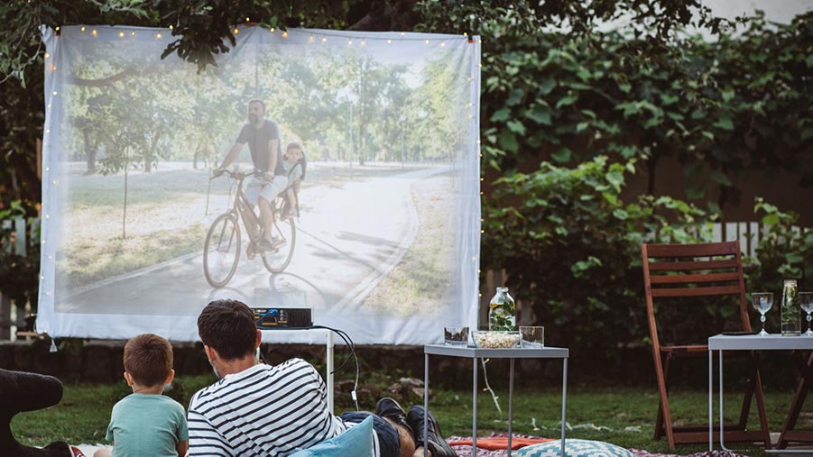 Father and son sitting outside on picnic blanket watching a projector movie in back yard with fairy lights.