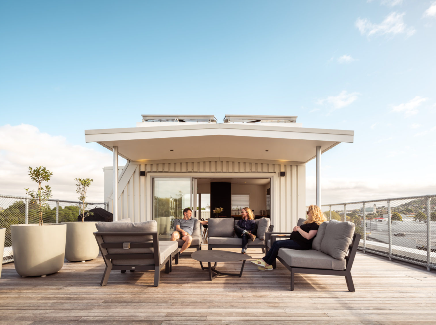 Three people sit on outdoor sofas chatting in a rooftop deck.