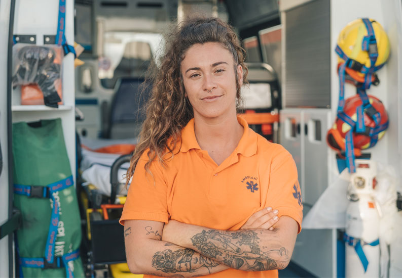 Female paramedic in NZ standing outside an ambulance with her arms crossed, looking at the camera.