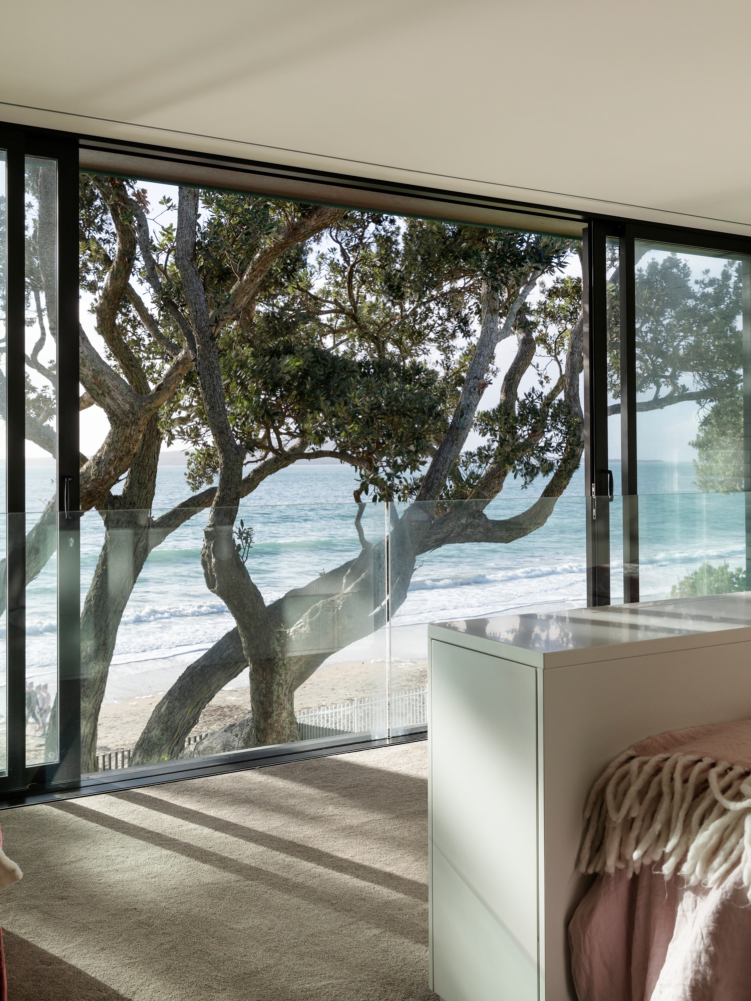 Interior view of a Hamptons-inspired home featuring floor-to-ceiling glass windows that provide an unobstructed view of a serene beachfront and mature trees. The natural light illuminates the room’s soft beige carpet and the corner of a white cabinet, with a glimpse of pink fabric, possibly from bedding or a garment. The blending of indoor comfort with the outdoor coastal landscape creates a tranquil and luxurious living space.