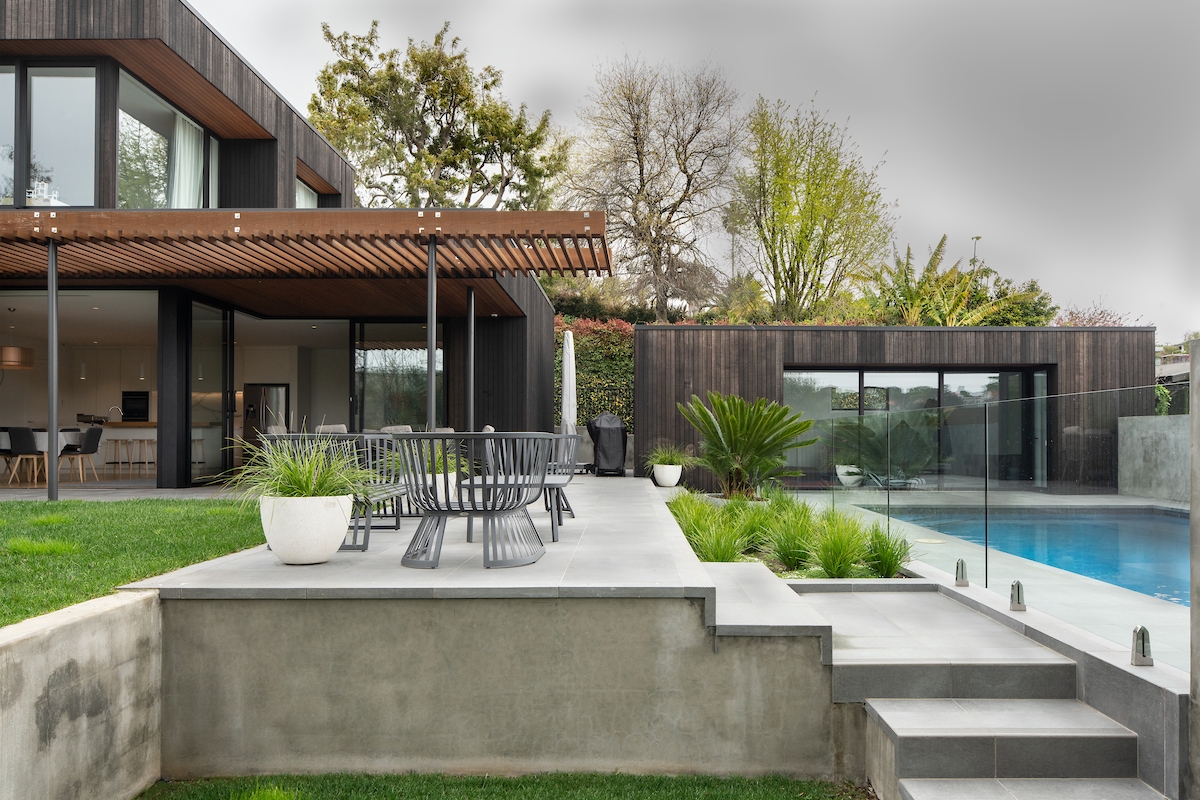 An image of a modern house with large glass windows and dark wooden panels. The house has a flat roof and is surrounded by greenery, including trees and shrubs. In the foreground, there is a concrete patio area with outdoor furniture, leading to a clear blue swimming pool enclosed by glass barriers. To the right of the pool, there’s another structure that mirrors the design of the main house. The sky is overcast, and the overall ambiance is serene and stylish.