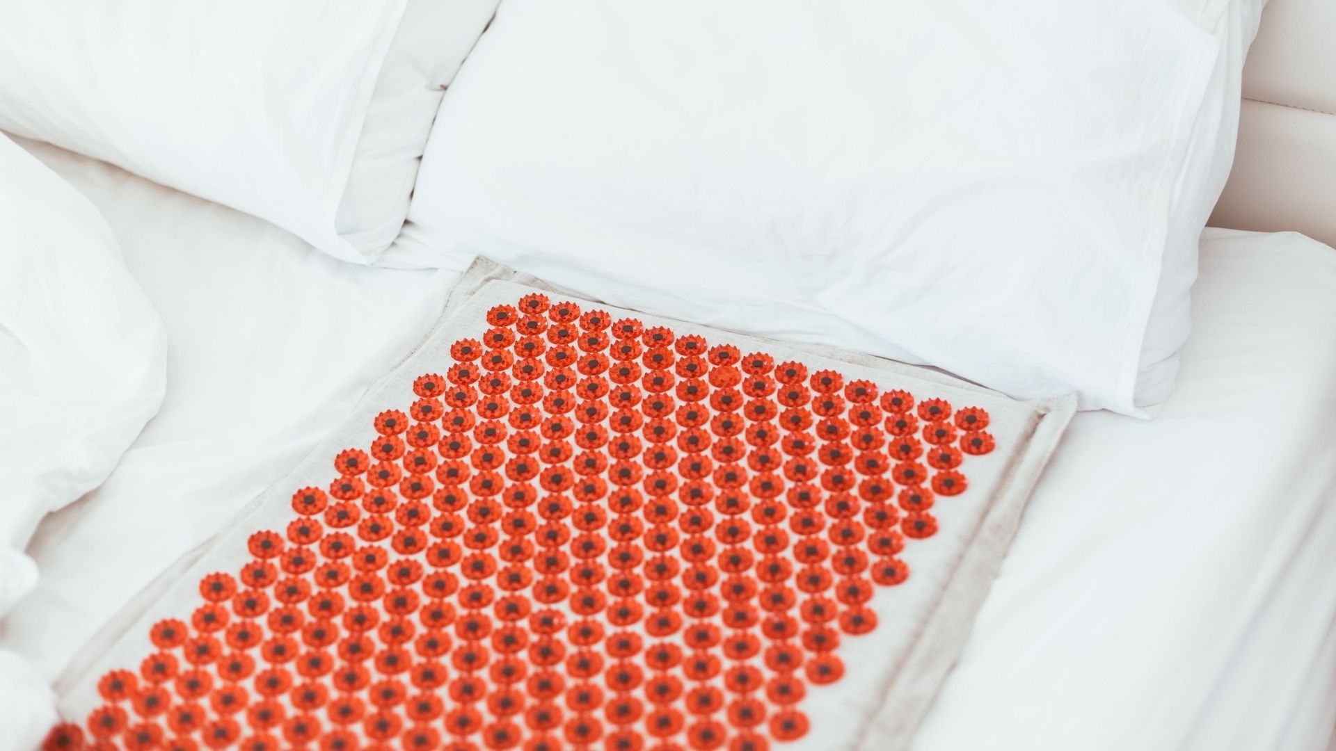 A grey and red acupressure mat is placed on a white bed, with a white pillow at the end of the mat.