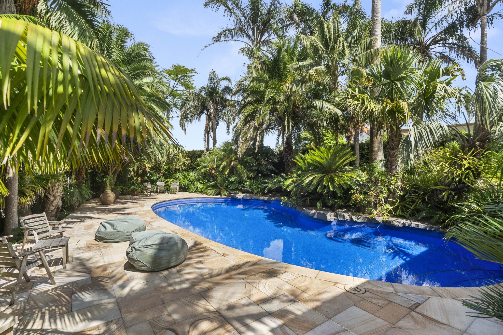 A backyard oasis with a blue swimming pool is surrounded by lush tropical gardens and trees. The area features poolside furniture and stone-tiled flooring.