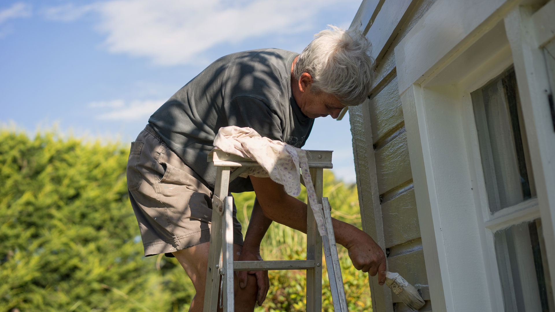 A man on a step ladder painting the outside of his NZ property in summer.