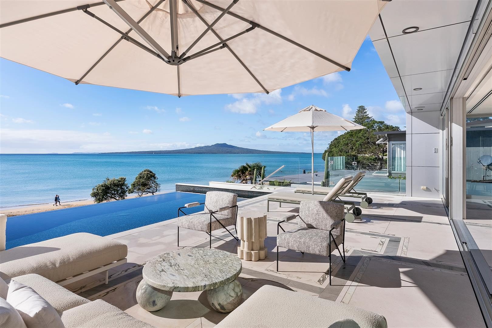 Luxury beachfront deck with infinity pool, shade umbrellas, and direct sandy beach access with Rangitoto views