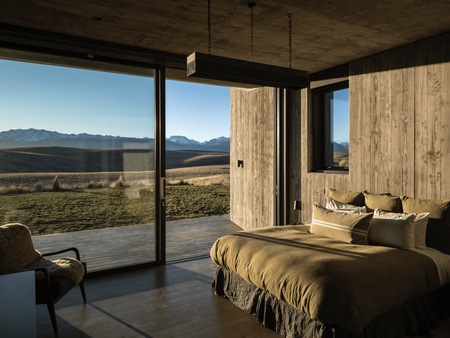 A master bedroom with large glass doors offering a panoramic view of rolling hills and distant mountains. The room features rustic wooden walls, a bed with earthy brown bedding, and minimalistic décor. Natural light streams in, casting warm shadows and enhancing the cozy, serene atmosphere of the space.