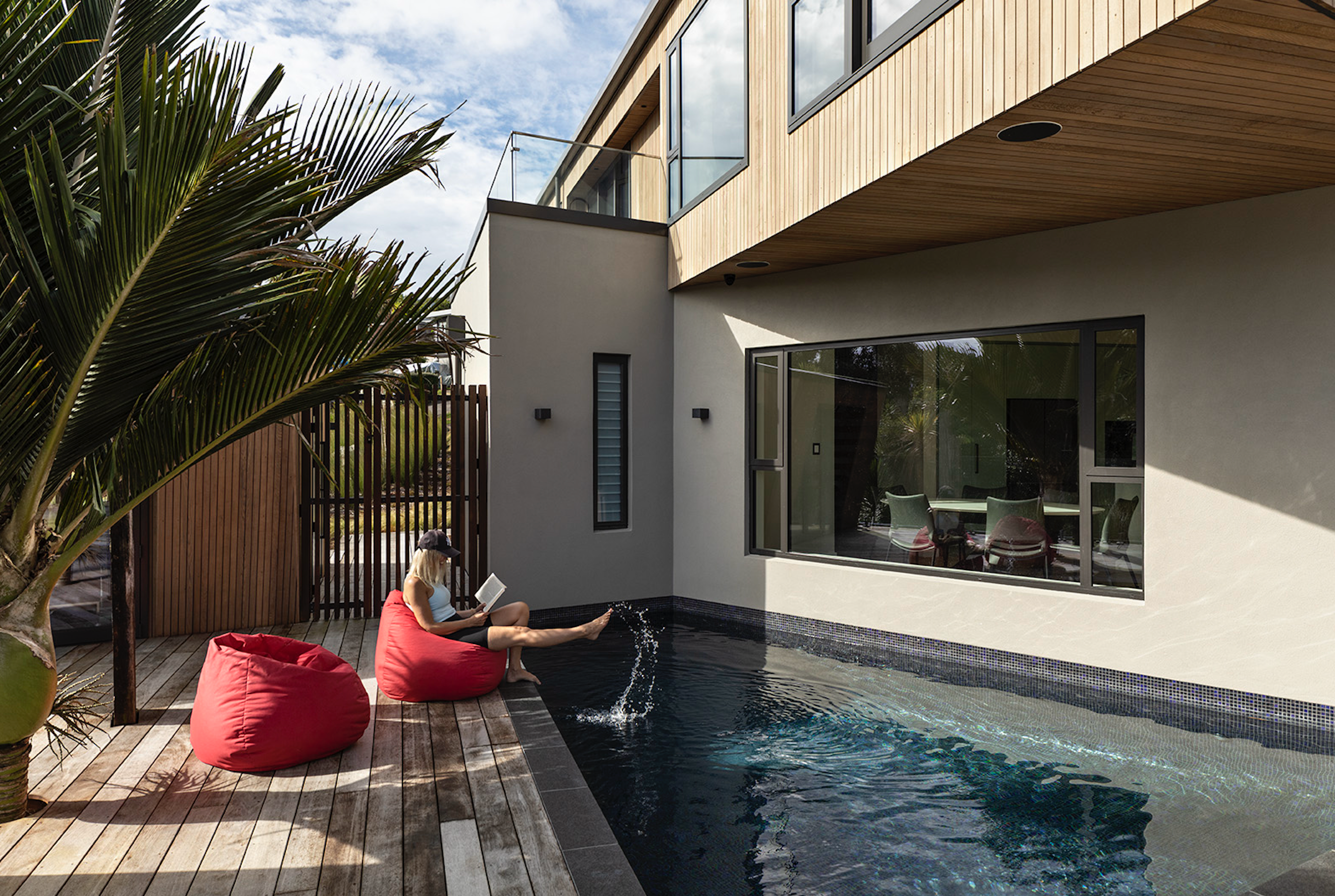 A modern backyard scene featuring a small rectangular in-ground swimming pool adjacent to a contemporary two-story house. A person is sitting on a red bean bag on the wooden deck, reading a book and dipping their feet into the pool. The house has large windows and a balcony above, with a palm tree partially visible in the foreground. The setting is cosy and intimate with a mix of sun and shade.