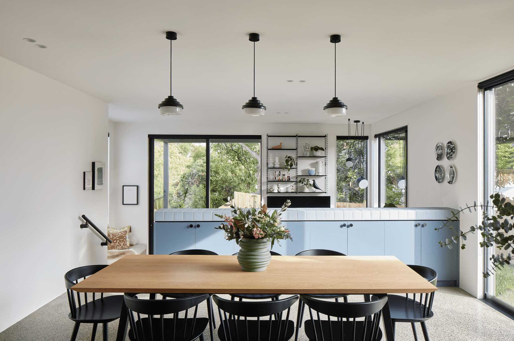 Bright, airy kitchen featuring a large wooden table with three pendant lights overhead, surrounded by white walls and blue cabinets, flooded with natural light.