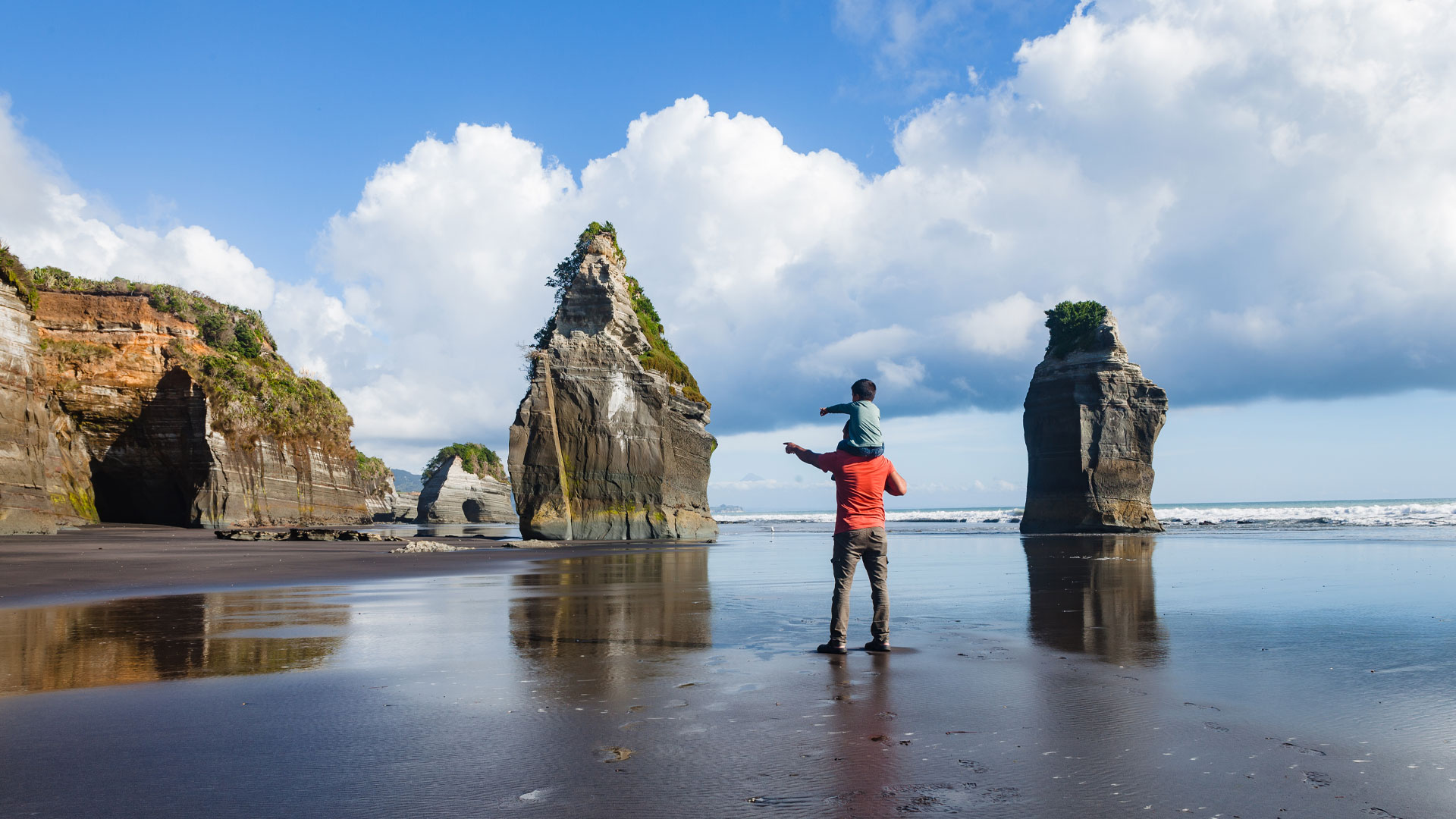 A father carrying a on his shoulders walking on the beach near the three sisters in Taranaki.