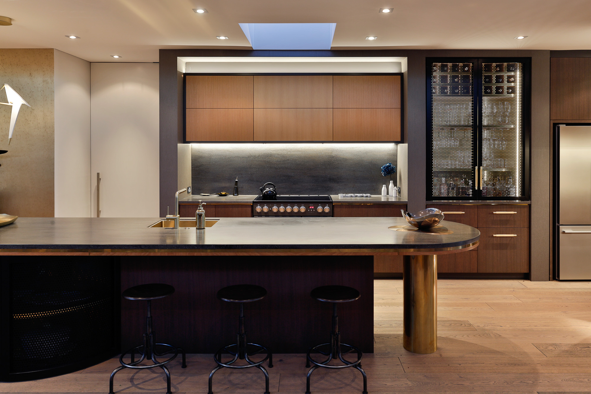 A modern kitchen featuring a large, curved island with seating for four. The splashback is a prominent feature, composed of a smooth, dark-toned material that spans the entire length of the counter and contrasts with the lighter countertop. Above the splashback is a row of sleek, white cabinets that match the ceiling design. The splashback’s uniform color and texture provide a backdrop for the stainless steel stove and built-in appliances, emphasizing their designs and creating an elegant and functional space.