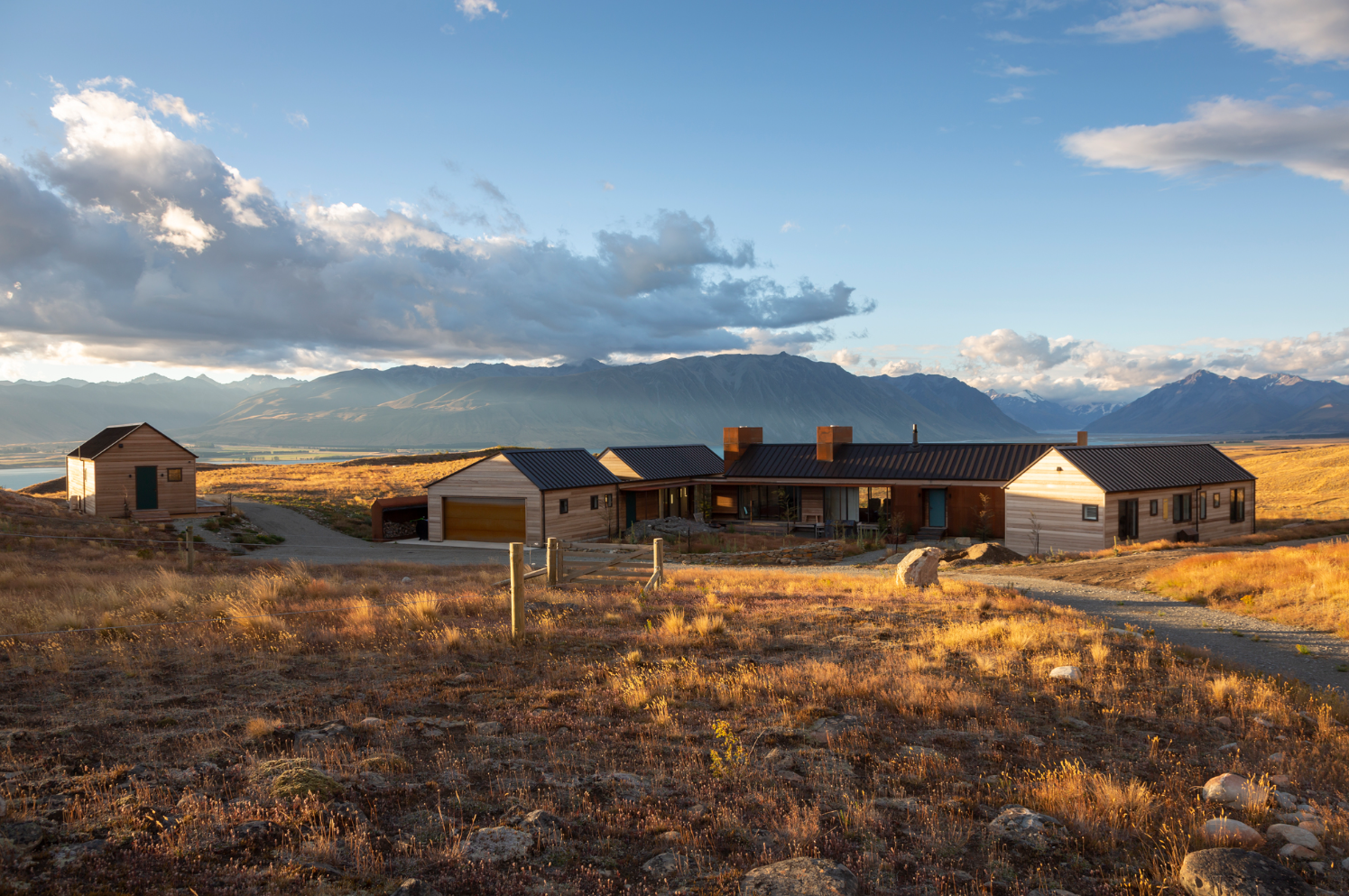 The image depicts a serene landscape during golden hour. It features a cluster of wooden buildings, residential structures and auxiliary buildings like garages or sheds, nestled amidst dry grassland. The terrain is covered in dry grass, giving it an autumnal feel, and there are scattered rocks on the ground. In the background, majestic mountains stretch across the horizon, partially obscured by clouds casting shadows upon them. The sky is partly cloudy with hues of blue and gold reflecting the setting or rising sun. Overall, it’s a tranquil scene capturing the beauty of nature and architecture co-existing harmoniously.