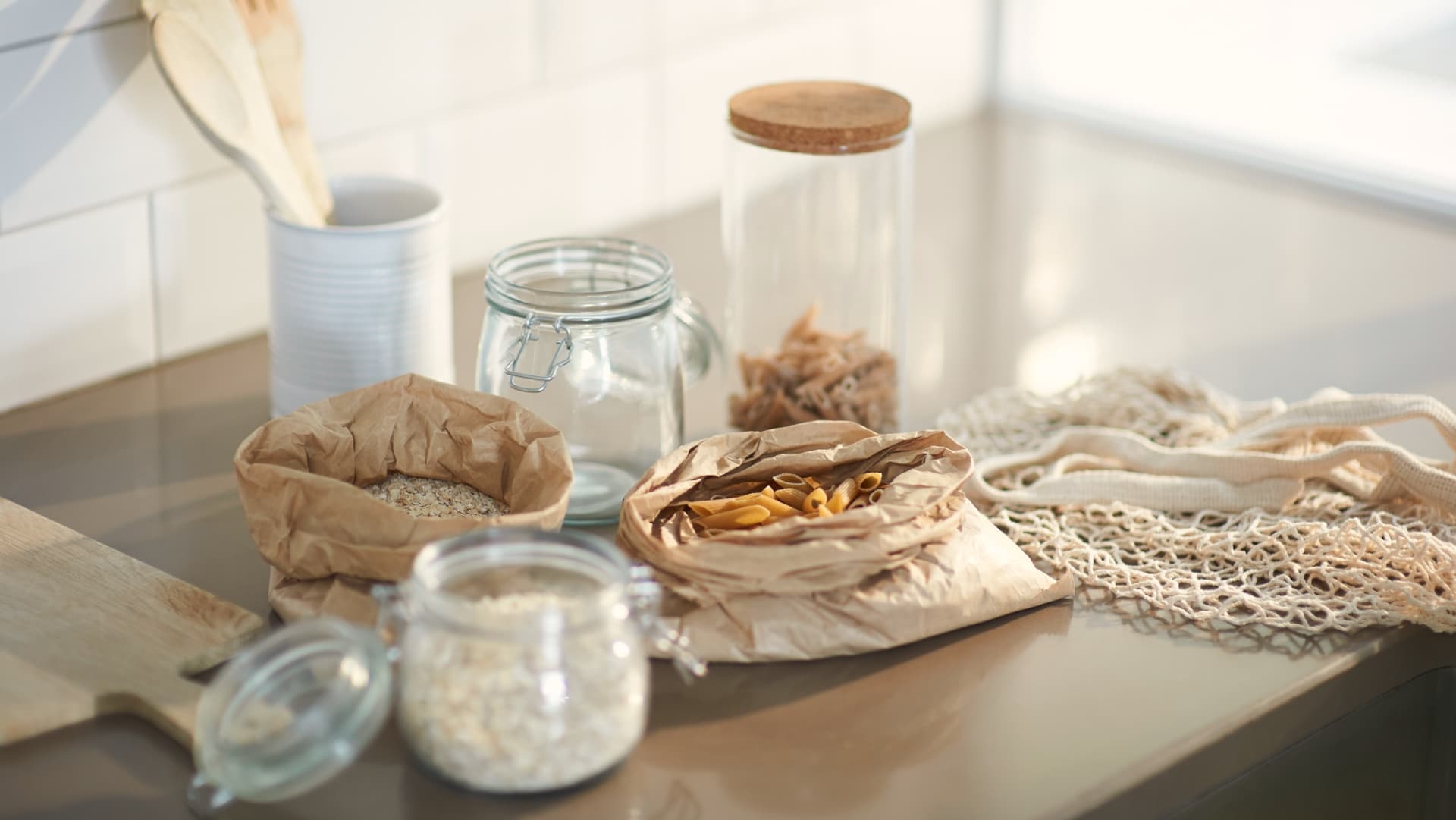 Stylized kitchen layout of pasta, rice and grains on bench being restocked into glass jars after weekly shop.