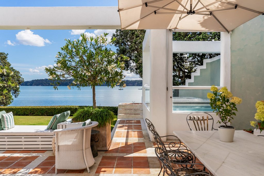 Waterfront terrace with cream pergola and shade sails, woven seating, manicured lawn, and direct Waitematā harbour views, with stairs leading to lower level