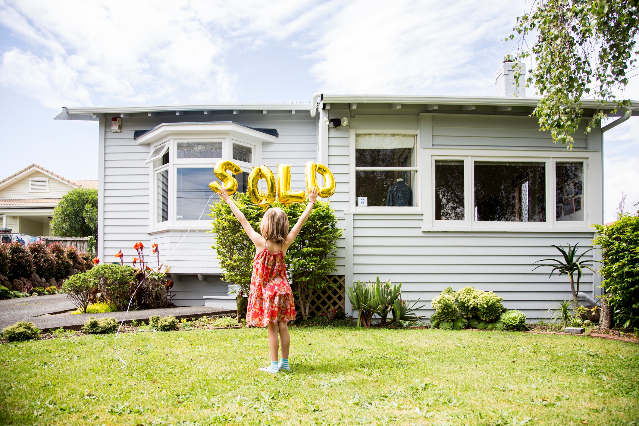 Little girl holds up gold balloons that read 'SOLD' in front of a house
