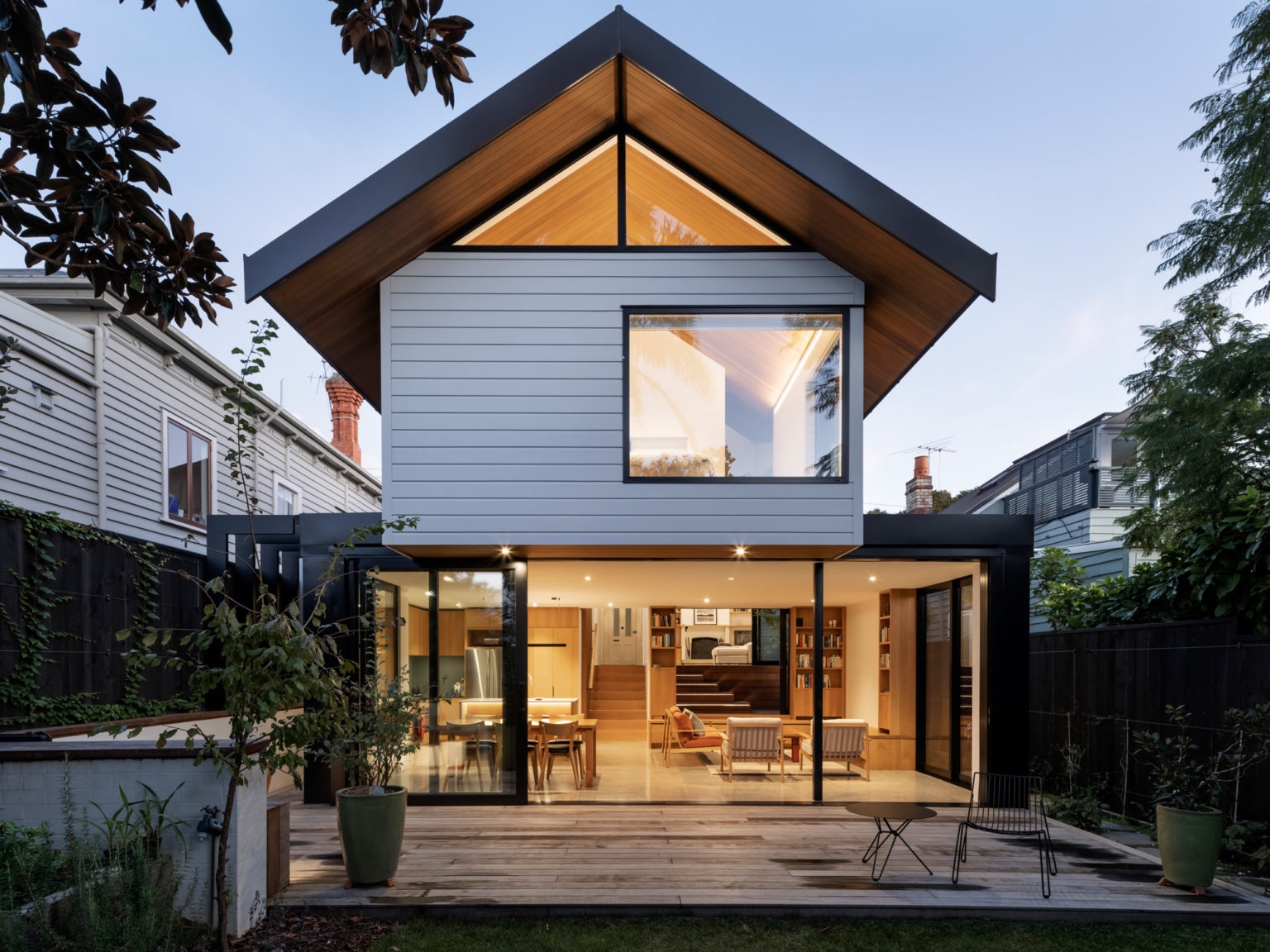 The image depicts a modern two-story house during twilight. The house features a combination of white siding and natural wood accents, with large windows that reveal the interior lighting, creating a warm and inviting atmosphere. The upper story has an asymmetrical gabled roof with a large triangular window that complements the geometric design. A well-manicured lawn surrounds the house, and there is a wooden deck at the forefront with outdoor furniture, suggesting a space for relaxation or entertainment. The sky is dimming, indicating either dawn or dusk, which adds to the serene ambiance of the setting. This architectural style might be relevant for real estate listings or discussions about home design.