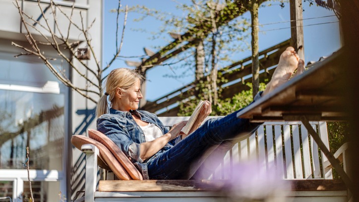 Woman sitting outside on wooden furniture. Her feet are up on the table and she is reading a book in the sun.
