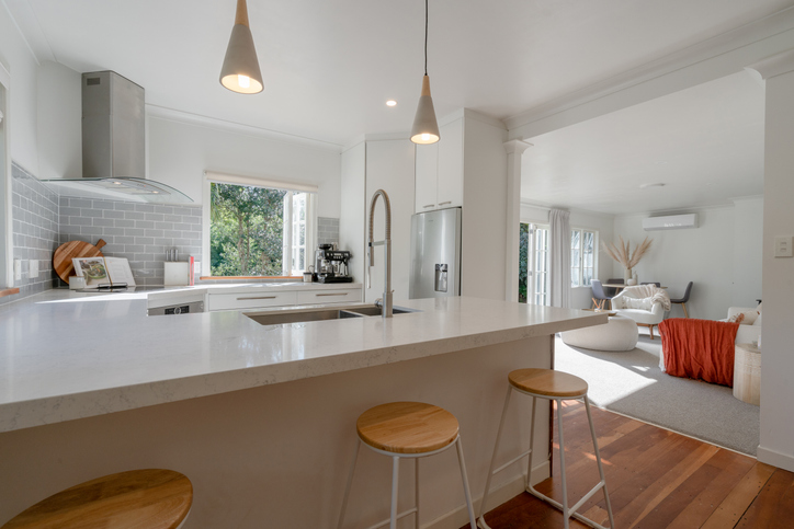A modern, light-filled open-plan living room and kitchen in a New Zealand home, featuring neutral tones, minimalist styling, and large windows that showcase a clean, high-value aesthetic.