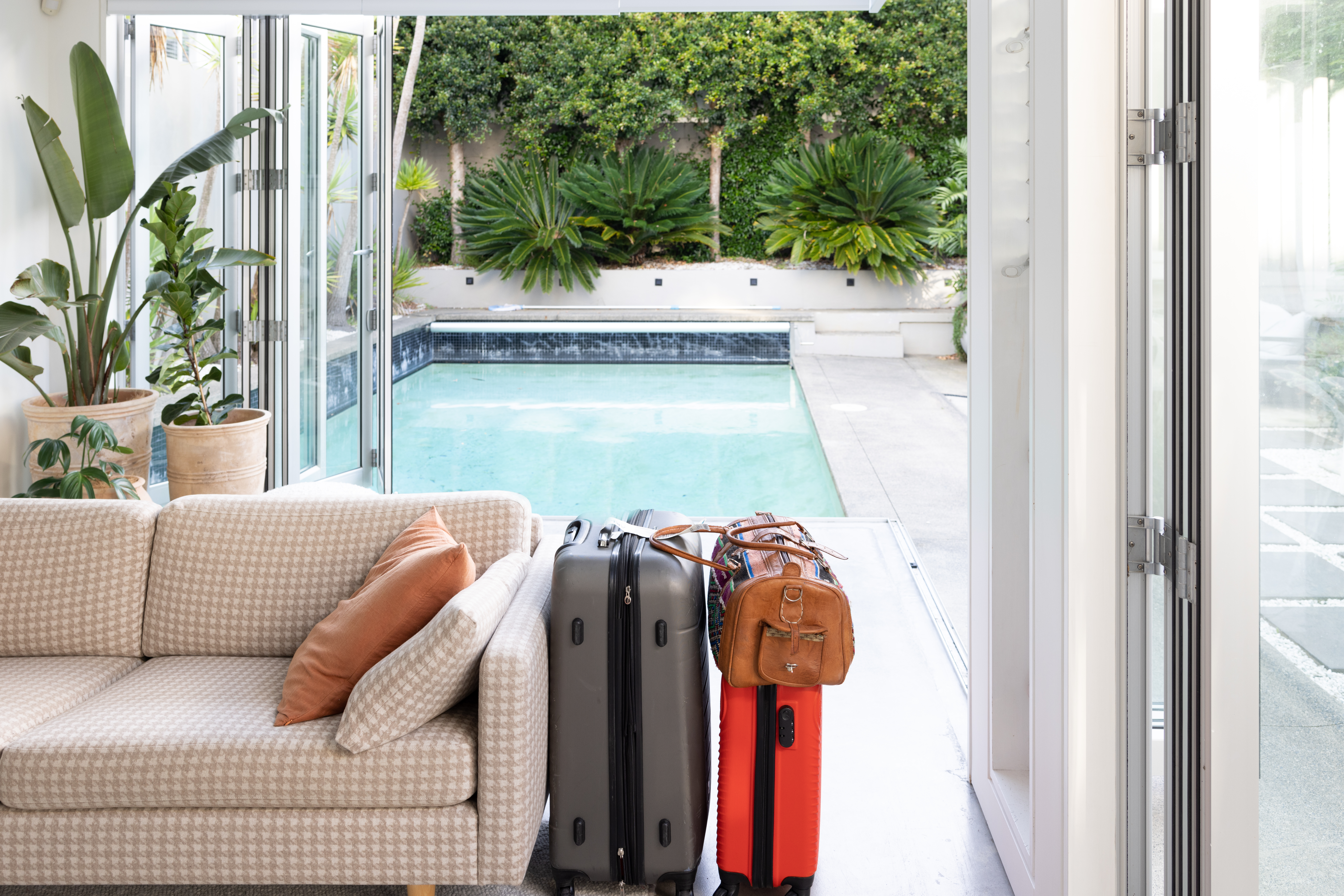 Two hard-shell suitcases placed inside a modern house, with large glass doors opening to a view of a sunlit swimming pool and outdoor lounge area. 
