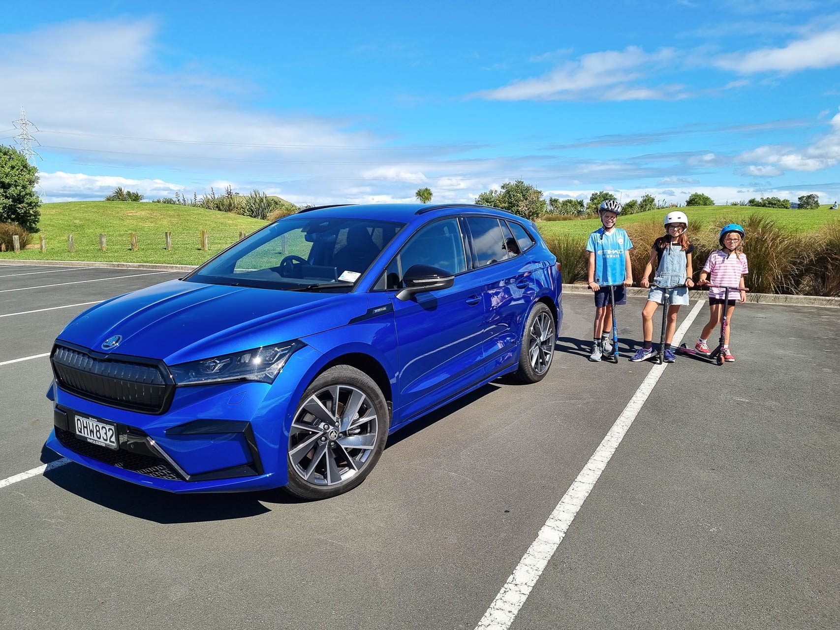 Blue Skoda Enyaq Sportline stationary shot from the front. Kids with scooter on the right.