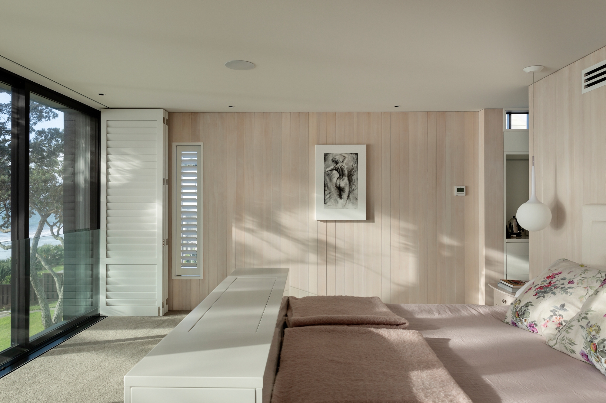 The image depicts a modern bedroom flooded with natural light. Light wood paneling adorns the walls and floor, creating a warm ambiance. A large bed with floral-patterned pillows sits at the center, flanked by white bedside tables. A framed black-and-white photograph of close-up flowers graces the opposite wall. An open door to the left reveals partial views of greenery outside. Overall, it’s a serene and inviting living space