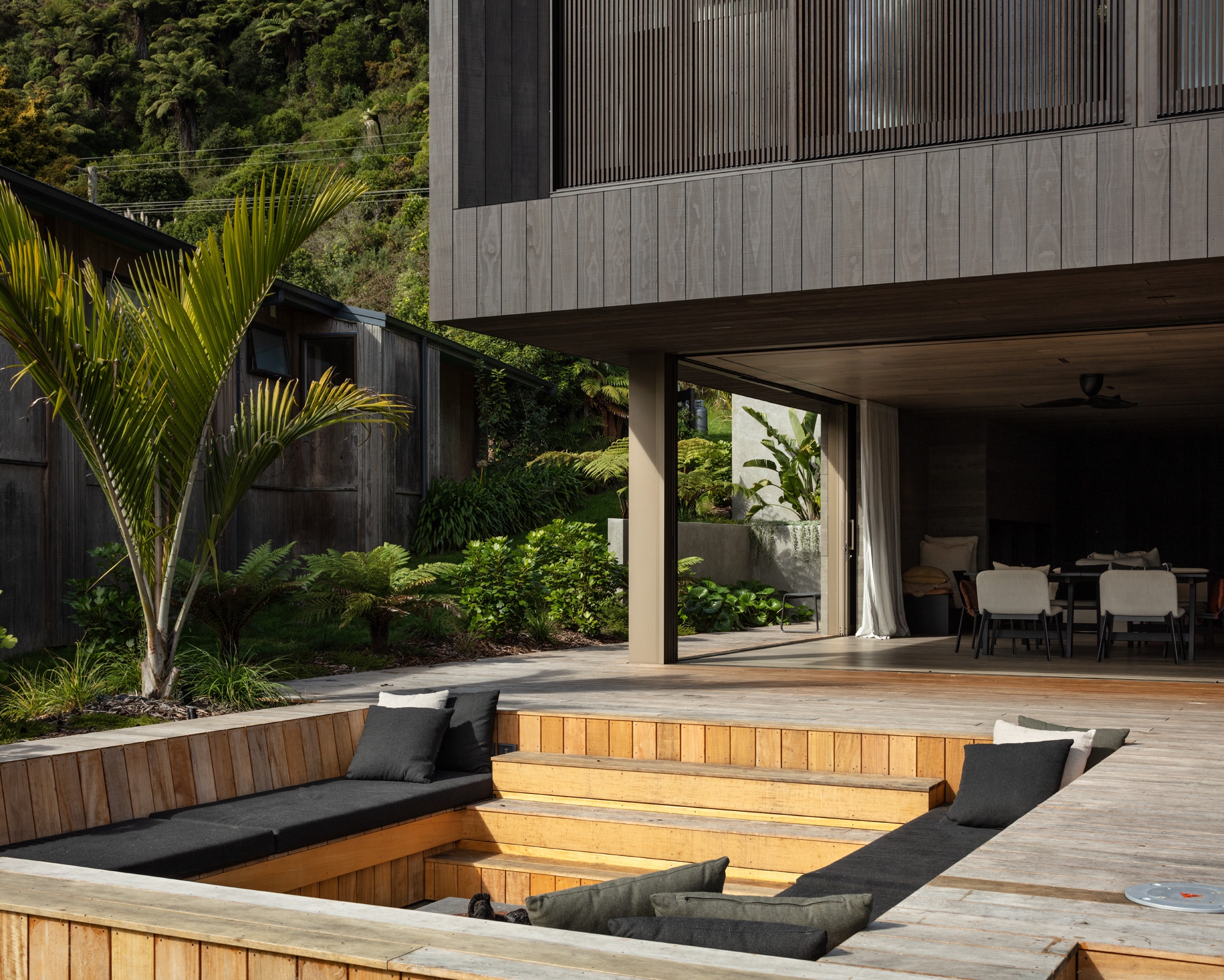 This image shows a modern outdoor living area with a sunken seating pit surrounded by a wooden deck. The seating area has wooden benches with dark grey cushions. The house has large glass doors that open to the deck, seamlessly blending indoor and outdoor spaces. The garden features lush greenery, including palm trees and various shrubs, creating a natural, tranquil environment. The combination of modern architecture and vibrant garden plants makes the space inviting and harmonious.