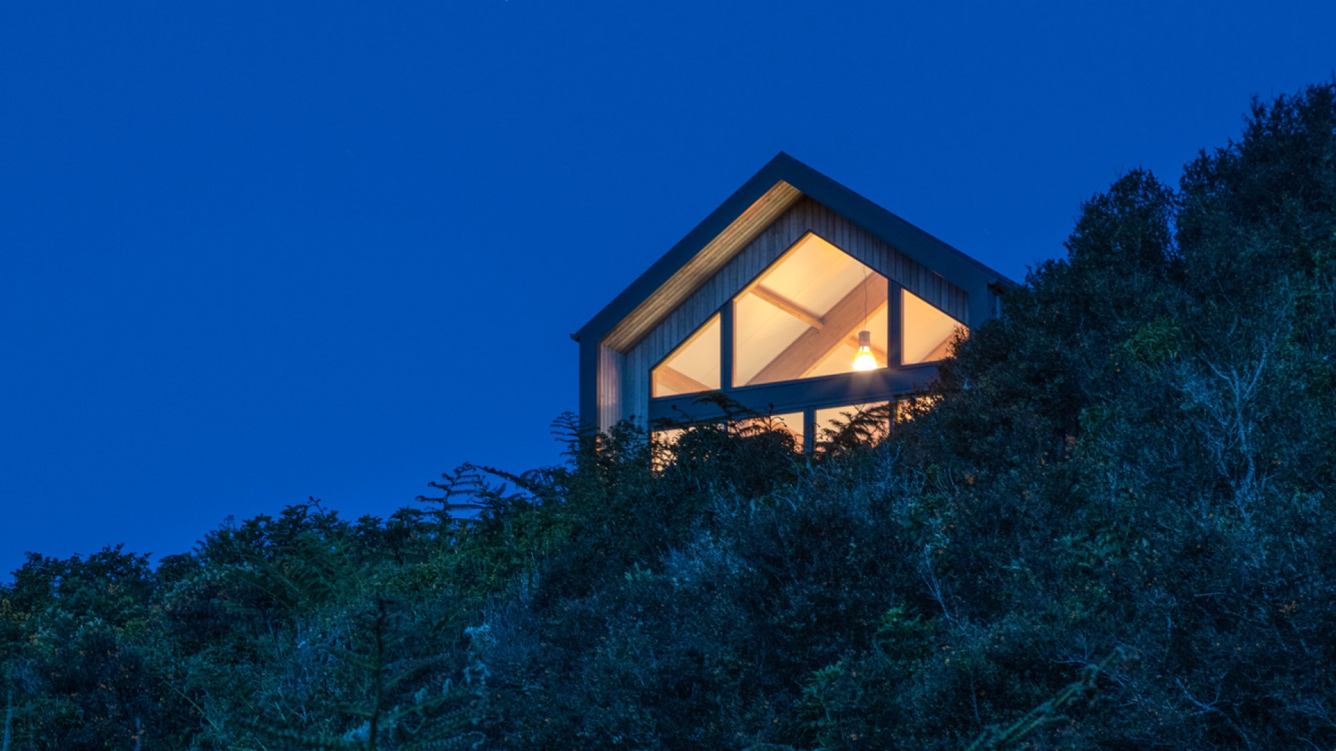 A modern house with large glass windows is perched on a hillside, illuminated from within against the twilight sky. The surrounding foliage is dense and dark, highlighting the warm light coming from a single visible interior lamp.