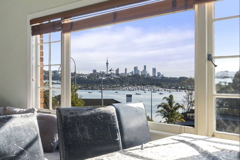 Windows frame a panoramic view of Auckland's skyline, the Sky Tower, and Waitemata Harbour dotted with boats. A worn chair sits in the foreground of a bright room.