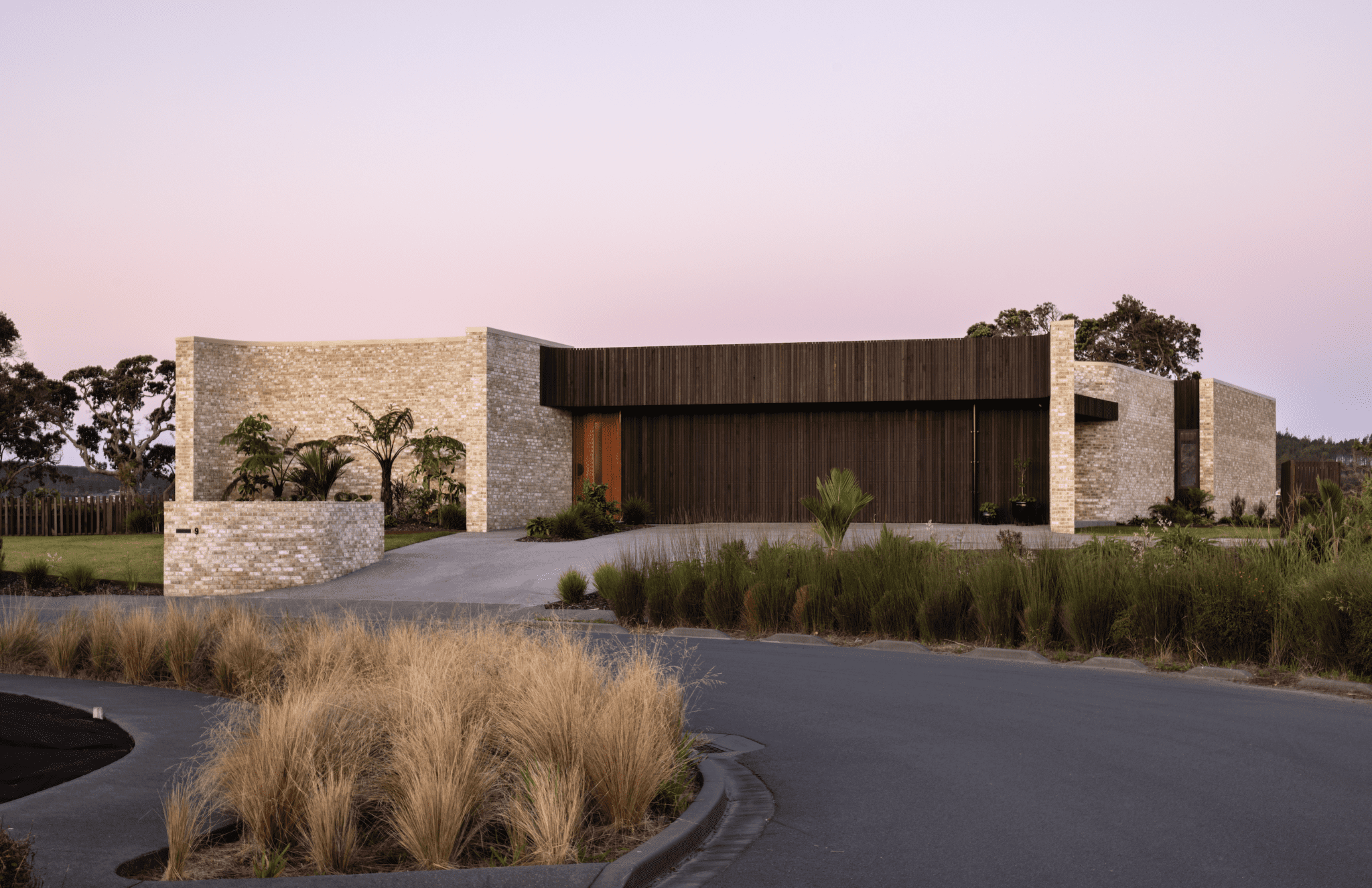 External shot of a white brick modern house with wooden panelled garage. The road side is surrounded by natural grasses leading the eye to the house.