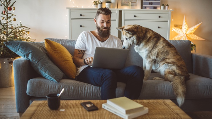 Man sitting on couch, using his computer. Husky sitting next to him on couch, looking at computer screen.