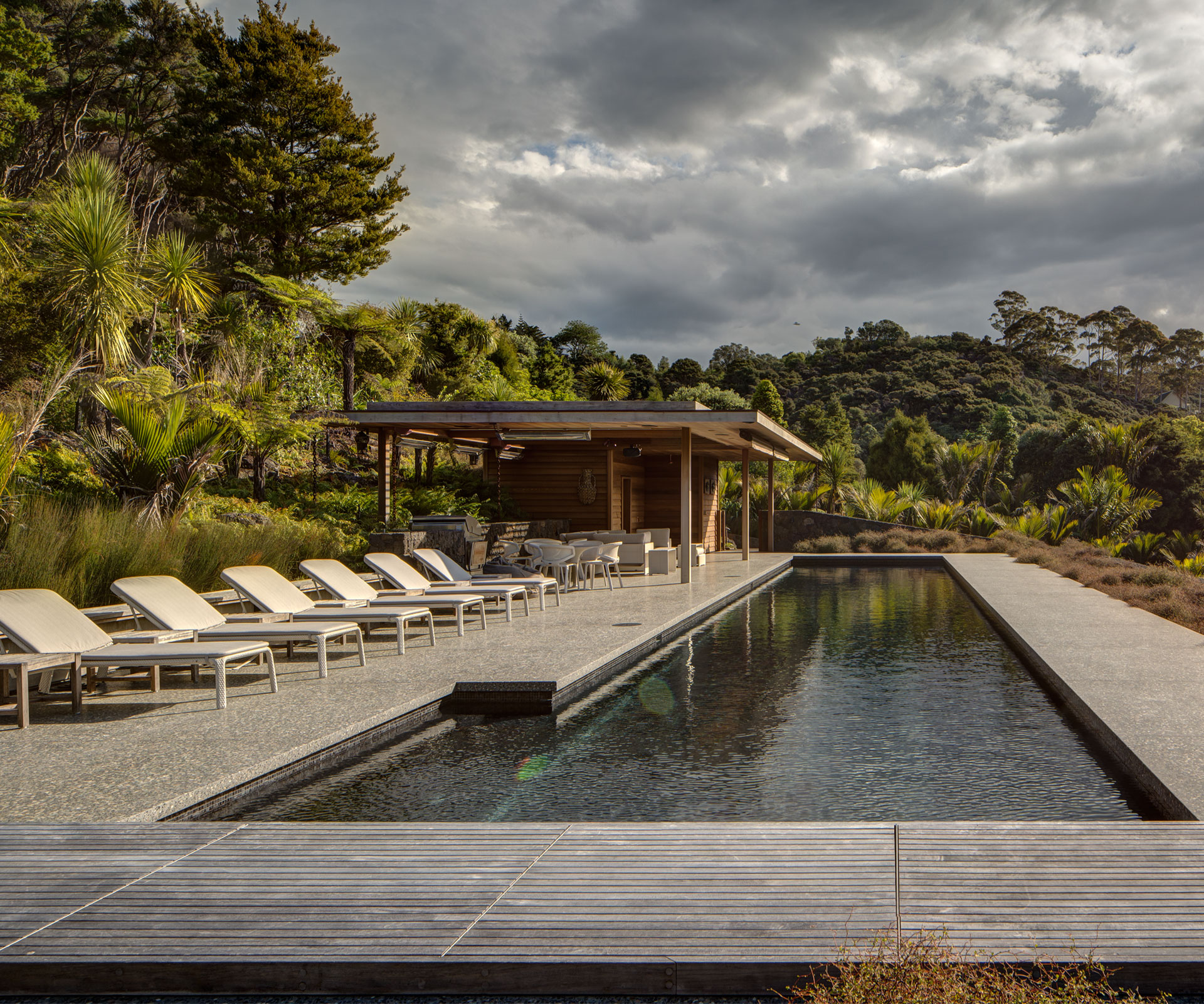 A serene backyard scene featuring a long, narrow in-ground swimming pool surrounded by lush greenery. Alongside the pool, there are several white lounge chairs and a covered wooden cabana equipped with dining furniture. The setting is tranquil with overcast skies and dense forested hills in the background.