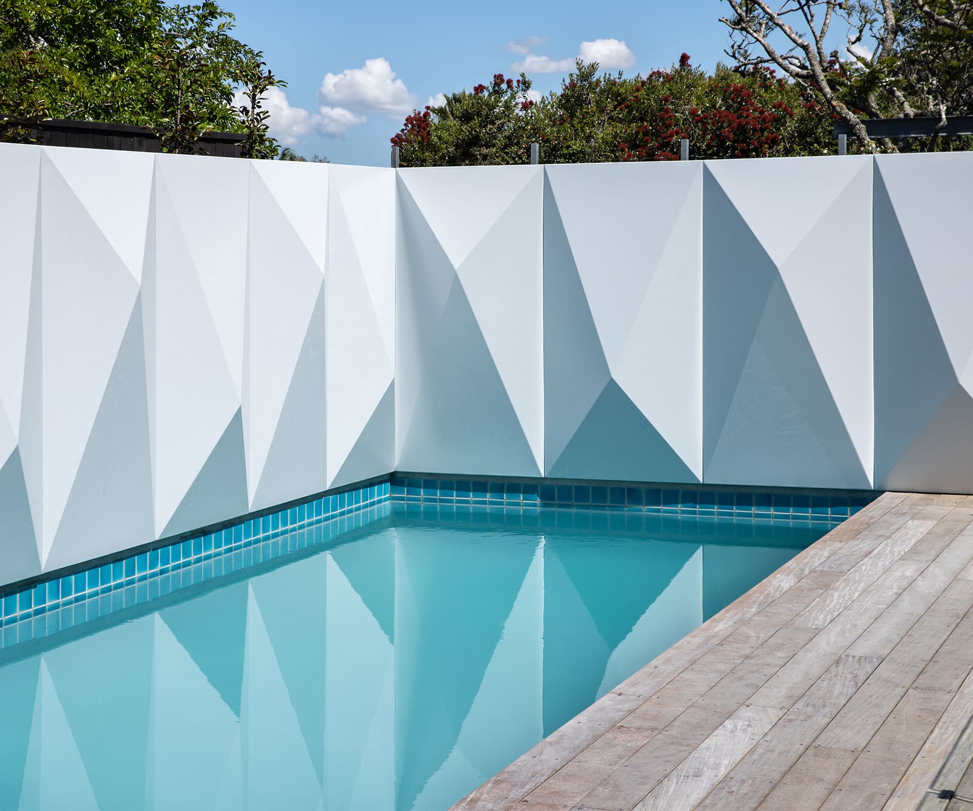 A modern swimming pool with clear blue water, surrounded by a wooden deck. The pool features a unique white geometric wall with triangular patterns, creating a striking visual effect. Lush green trees and a clear blue sky are visible in the background, enhancing the serene and contemporary ambiance.