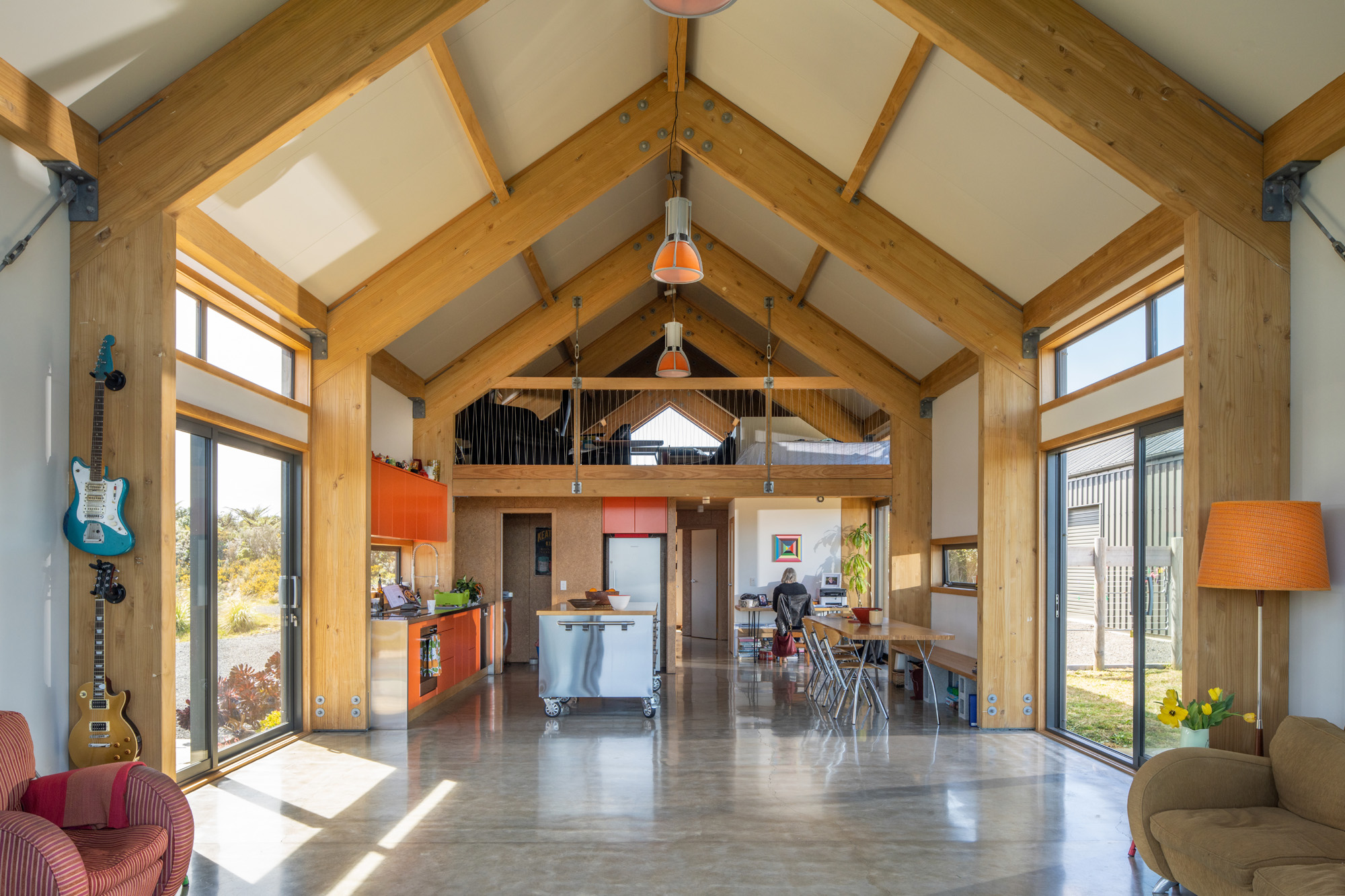 The spacious and well-lit interior of a modern house features high ceilings with exposed wooden beams. Natural light floods the room through large windows and glass doors. On the left, a wall-mounted electric guitar stands next to a red-patterned armchair. In the center, an open kitchen area boasts orange cabinetry, stainless steel appliances, and a central island with stools. To the right of the kitchen, a dining area is set with a table for four. Above this space, an open loft accessible by stairs adds an interesting architectural element. The polished concrete flooring completes the contemporary design.