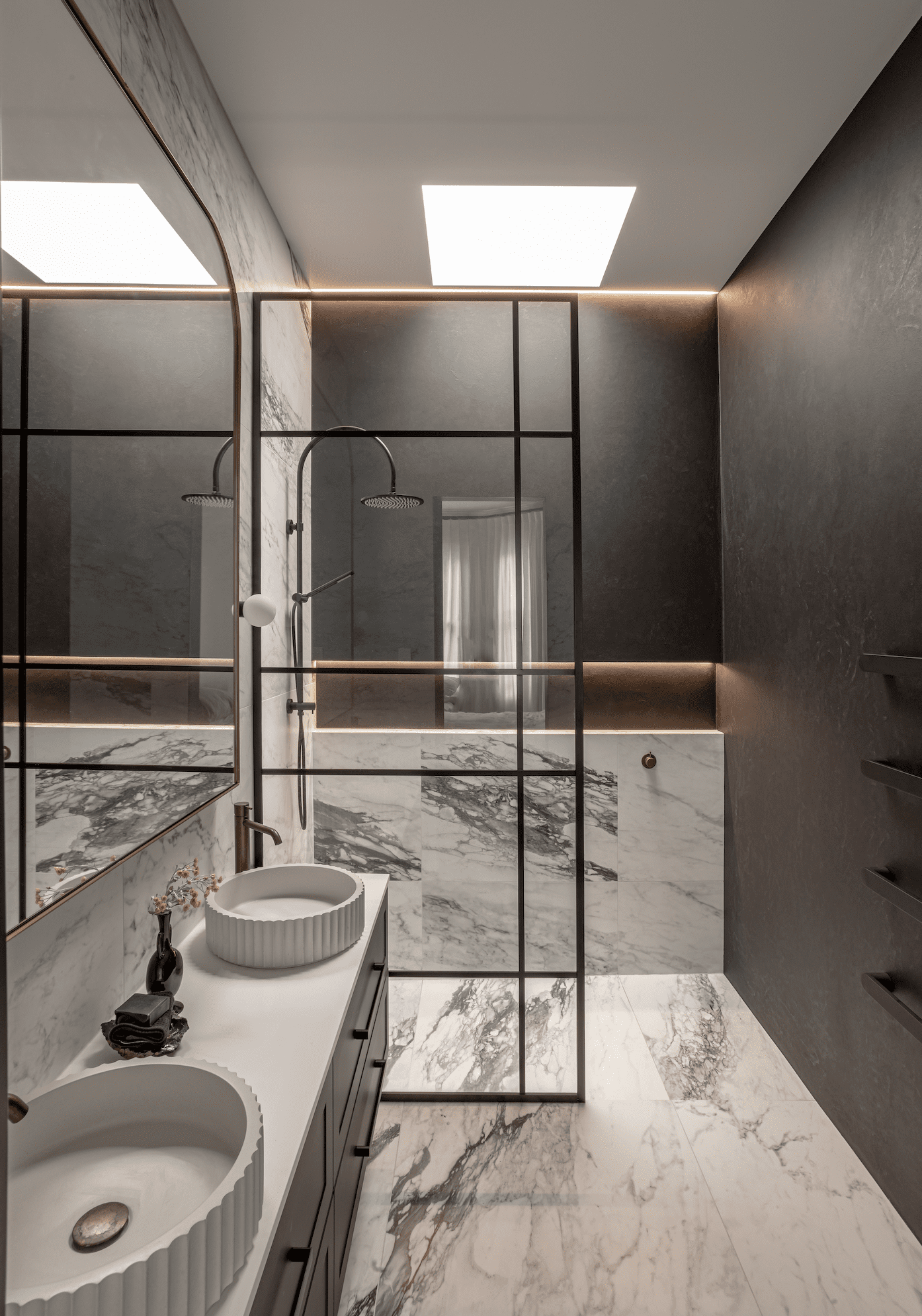 Luxurious modern bathroom with a marble theme, featuring a double vanity with textured round vessel sinks, sleek black fixtures, and a glass shower enclosure with black grid frames. The room is highlighted with soft, recessed lighting and a skylight above, creating a warm and elegant atmosphere.