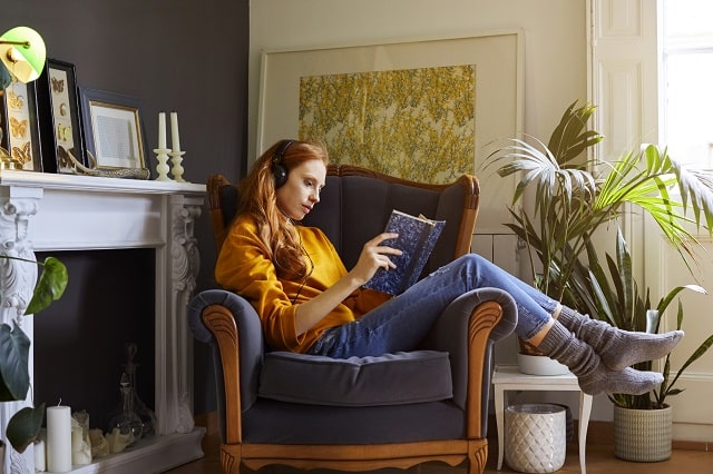 Woman wearing a brightly coloured jumper reading her book in an armchair by the fireplace.