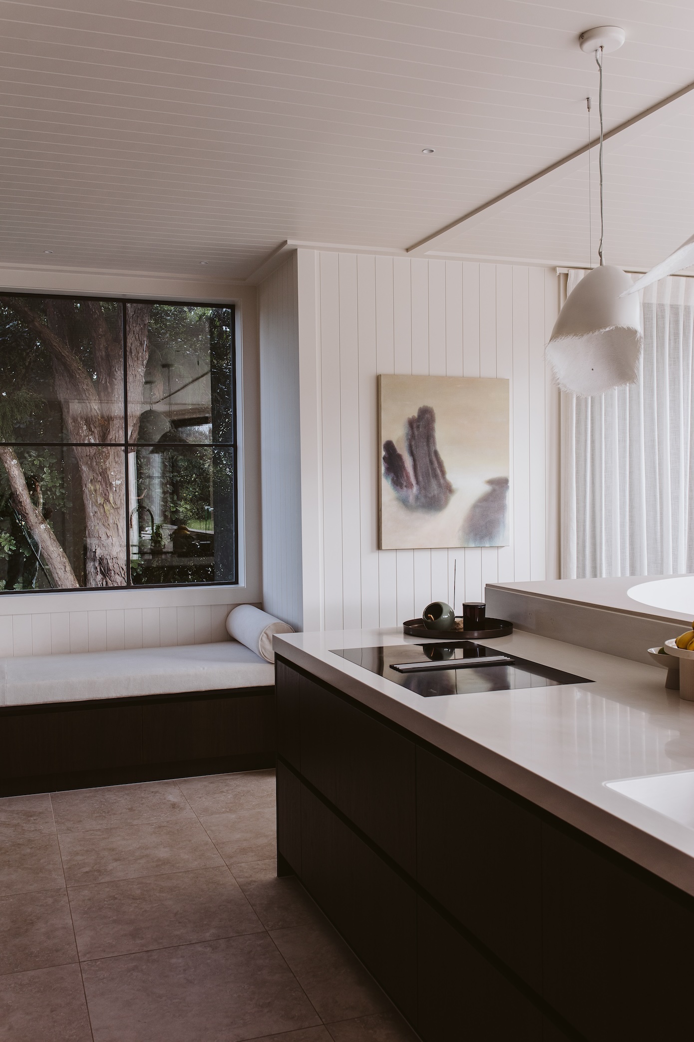 The kitchen is crisp-smart with a Corian Cirrus White by APT benchtop and Stardust Walnut Shinnoki Veneer by Vidaspace cabinetry. The benchseat has a view of rimu in a protected reserve. Glare by Kyla Covic hangs between the kitchen and dining.