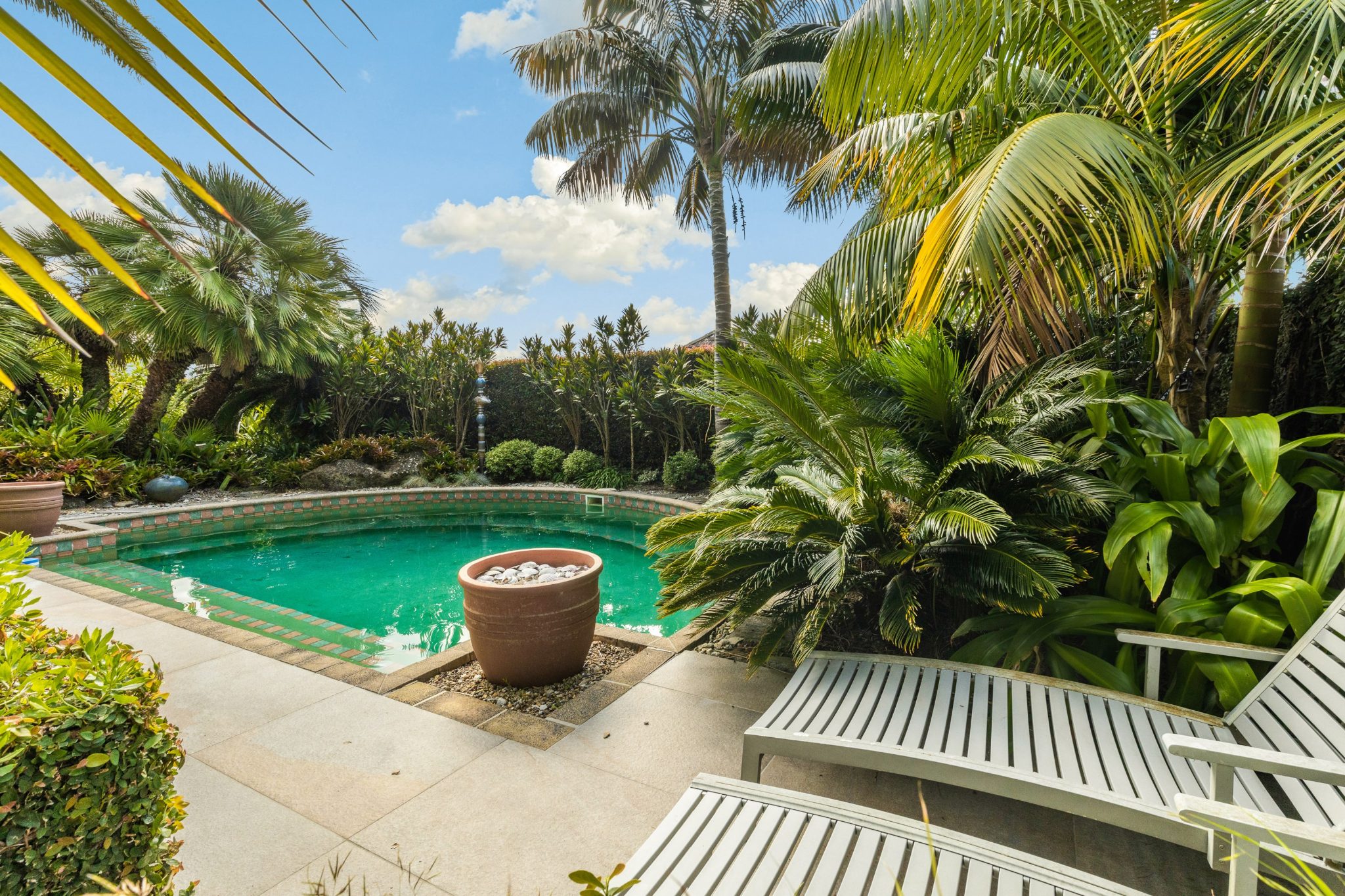 The image shows a kidney-shaped swimming pool surrounded by lush greenery. Turquoise water contrasts with a brown circular planter containing floating plants at the pool’s center. Tropical palm trees and other plants create a private oasis. Grey lounge chairs rest on square stone tiles by the poolside. The partly cloudy sky suggests a warm climate.