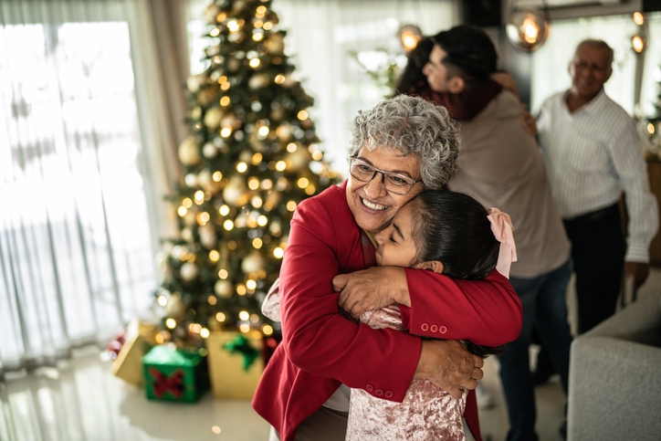 Family gather around the Christmas tree as Grandma embraces grandchild in a hug.