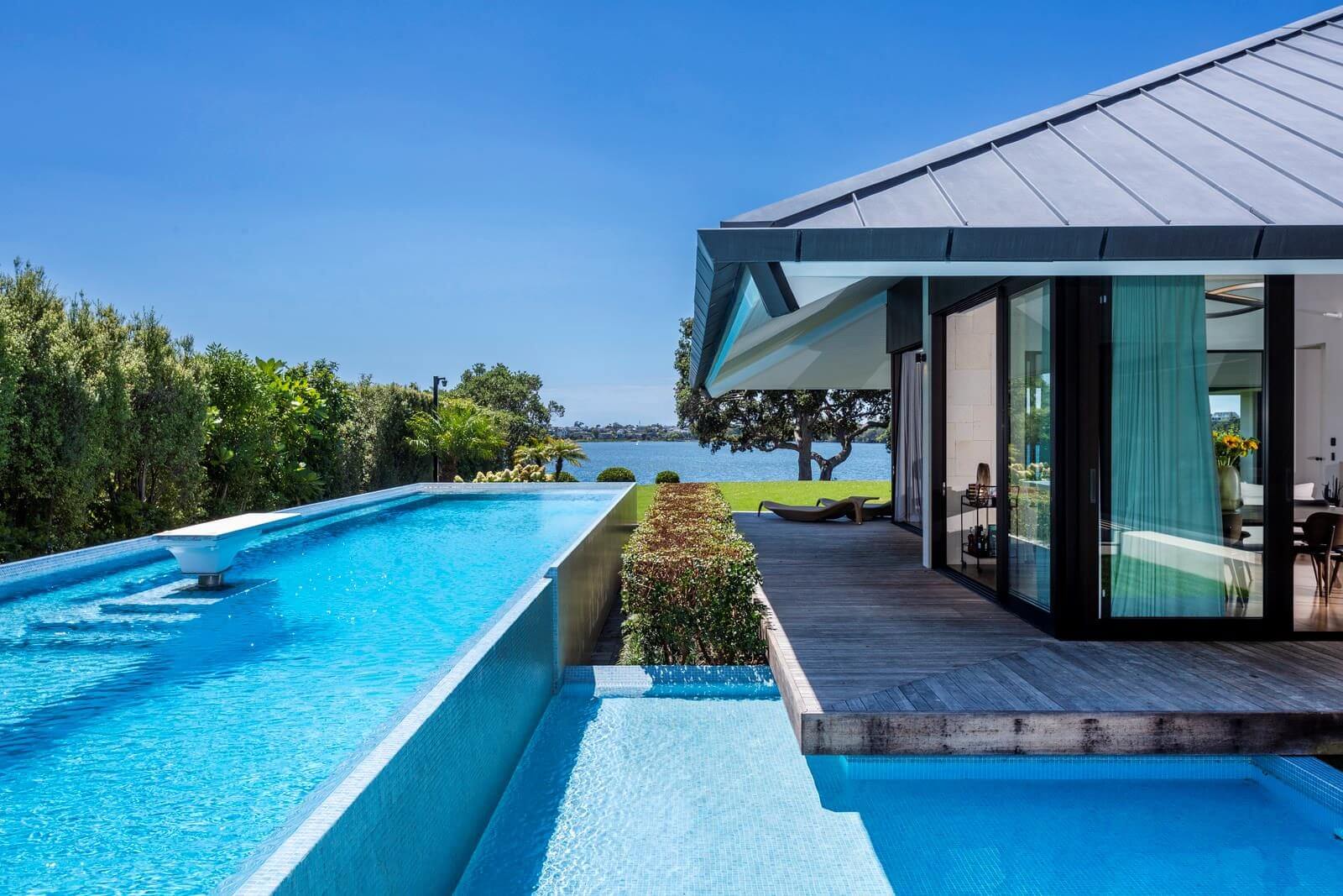 Modern lakefront pavilion with bright blue infinity pool, dark timber deck, and Lake Pupike views beyond lush gardens