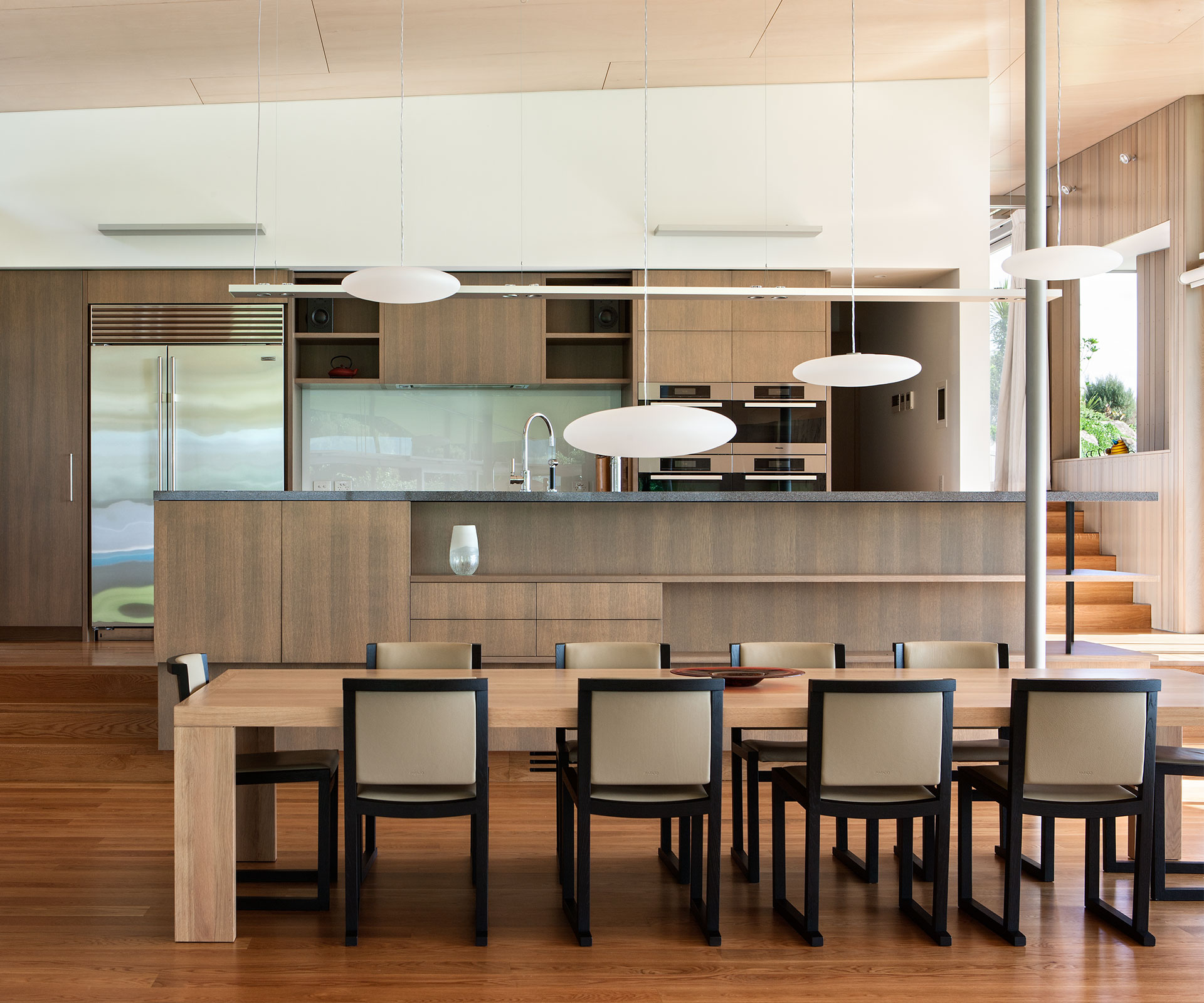 Modern kitchen with wood cabinetry, stainless steel refrigerator, pendant lights above the island, and a glass splashback.