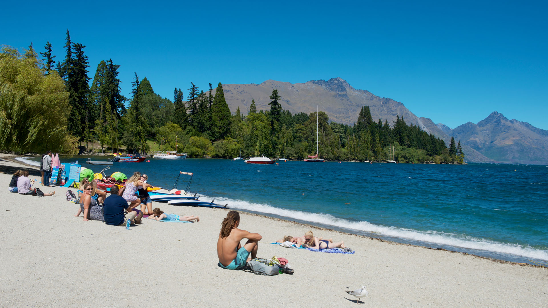 A photo of the beachfront at Lake Wakatipu, Queenstown, with people relaxing in the sun.