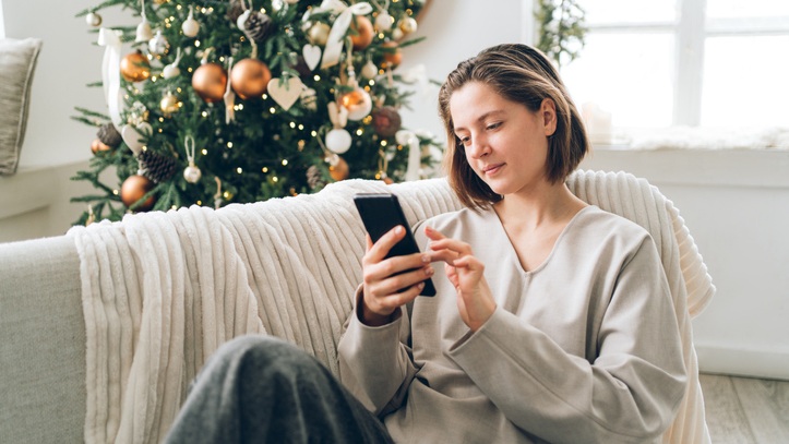 Woman sitting on couch shopping on her phone. There is a Christmas tree in the background.