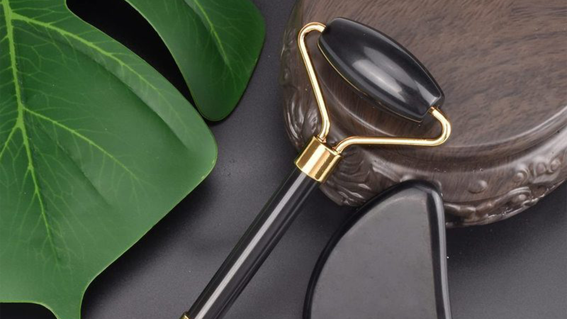 Black with gold trim crystal facial roller placed on brown wooden dish and next to a monstera leaf.