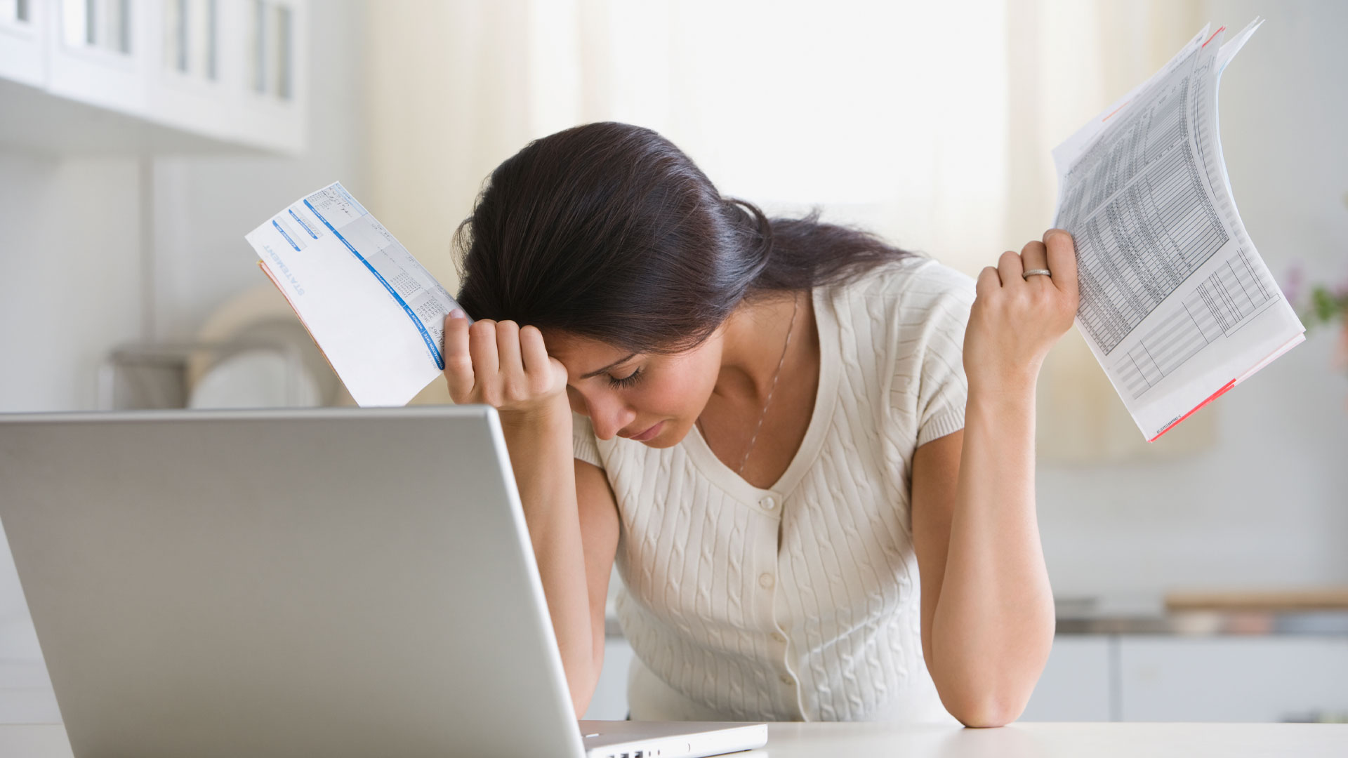 Stressed out woman holding financial documents and looking at homes for sale in NZ on her laptop.