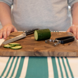 Half sliced cucumber sitting on a cutting board with a knife and a peeler