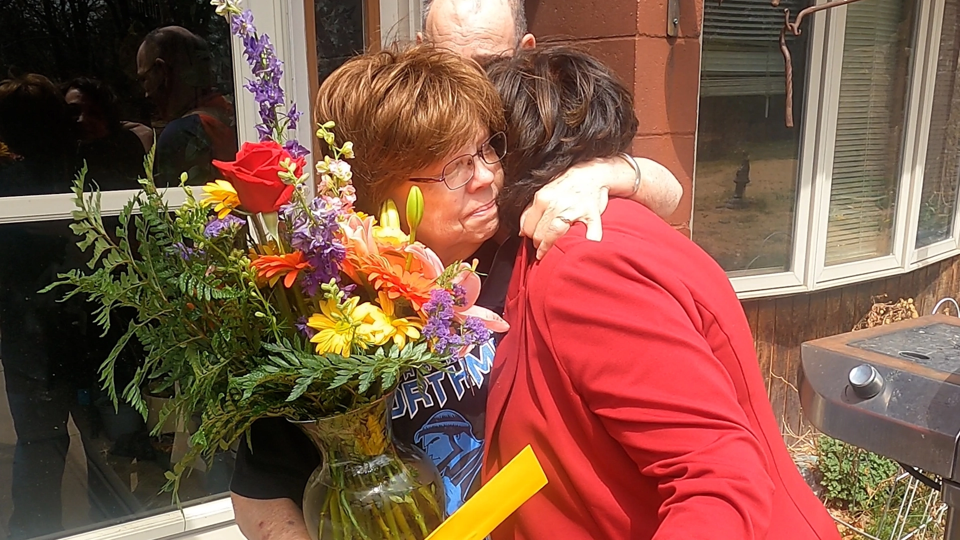 Woman hugging an older woman who is holding a vase of flowers and yellow envelope outside