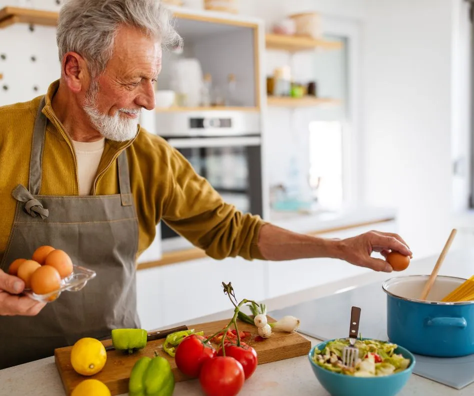 Older man with gray hair wearing a mustard sweater and gray apron cooking in a kitchen with eggs and vegetables