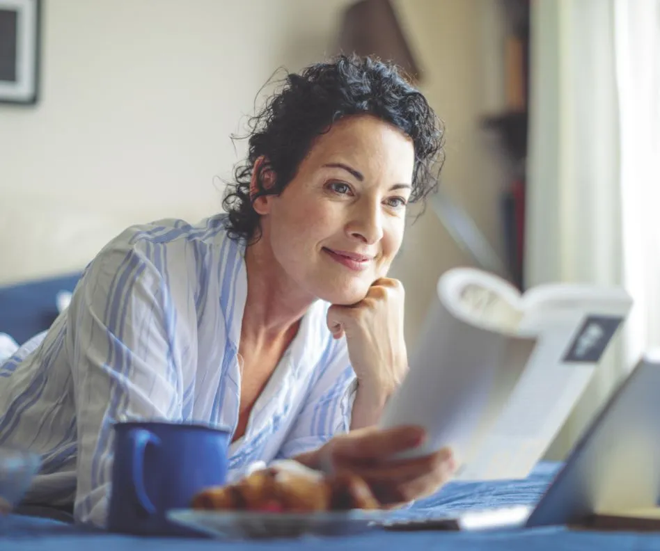 Middle age woman with dark brown hair smiling and relaxing while laying down reading a book with blue coffee mug