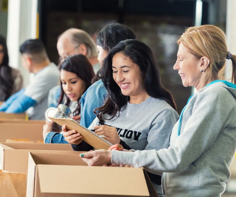 Group of people in gray and blue clothing volunteering and filing boxes with goods while smiling