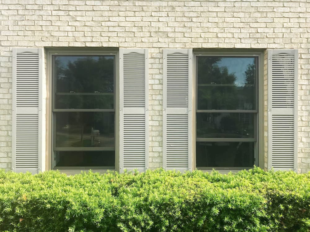 Two new wood double-hung windows with beige shutters on an ivory brick home