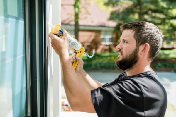 a man reapplying sealant along the exterior of his house near a window